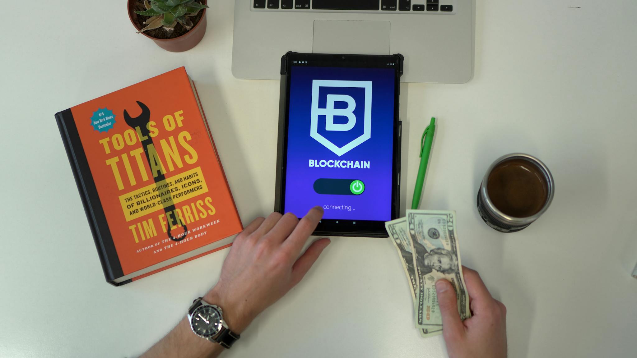 Flat lay image featuring a tablet with blockchain app, US dollars, and a book on a desk.